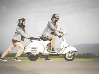 Seorang pemuda duduk di atas Vespa berwarna putih, sementara seorang wanita mendorongnya di jalanan kota yang tenang