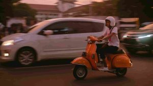 Seorang pria mengenakan helm mengendarai Vespa berwarna biru di jalan kota yang ramai dengan mobil